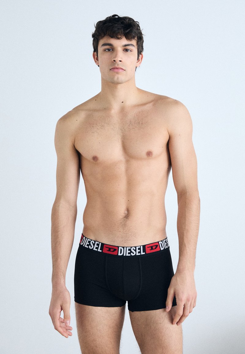 Boxer noirs avec une ceinture logo "DIESEL" rouge et blanche, présentant un design ajusté et une texture de tissu douce, modèle se tenant contre un fond uni.