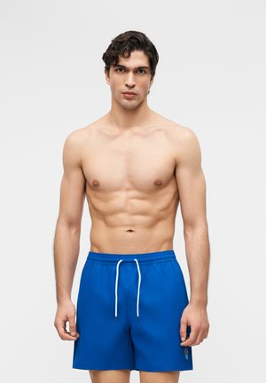 Shirtless young man with curly dark hair wearing blue swim shorts with white drawstrings, standing against a plain light background.