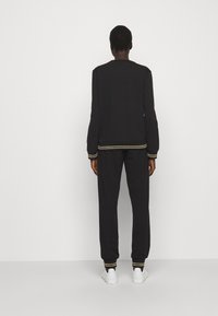 Sudadera negra y pantalones con puños de rayas doradas. La tela parece suave. El modelo se encuentra de espaldas contra un fondo blanco liso.