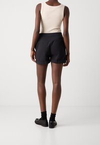 Pantalones cortos negros con una textura suave y dobladillos enrollados, combinados con una camiseta sin mangas color beige. Modelo luciendo zapatos negros con correas y calcetines.