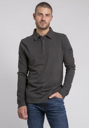 Homme aux cheveux courts portant un polo à manches longues gris foncé et un jean bleu, debout avec une main dans la poche devant un fond uni.