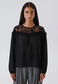 Donna con lunghi capelli scuri indossa una blusa nera trasparente con yoke in pizzo floreale e volant, abbinata a pantaloni grigi plissettati su uno sfondo semplice.