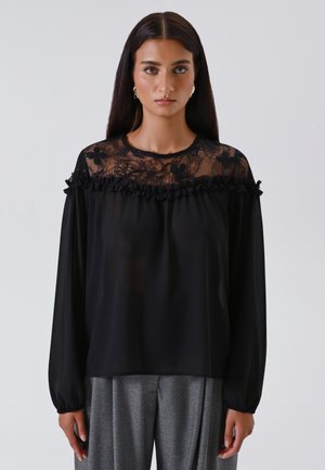 Donna con lunghi capelli scuri indossa una blusa nera trasparente con yoke in pizzo floreale e volant, abbinata a pantaloni grigi plissettati su uno sfondo semplice.