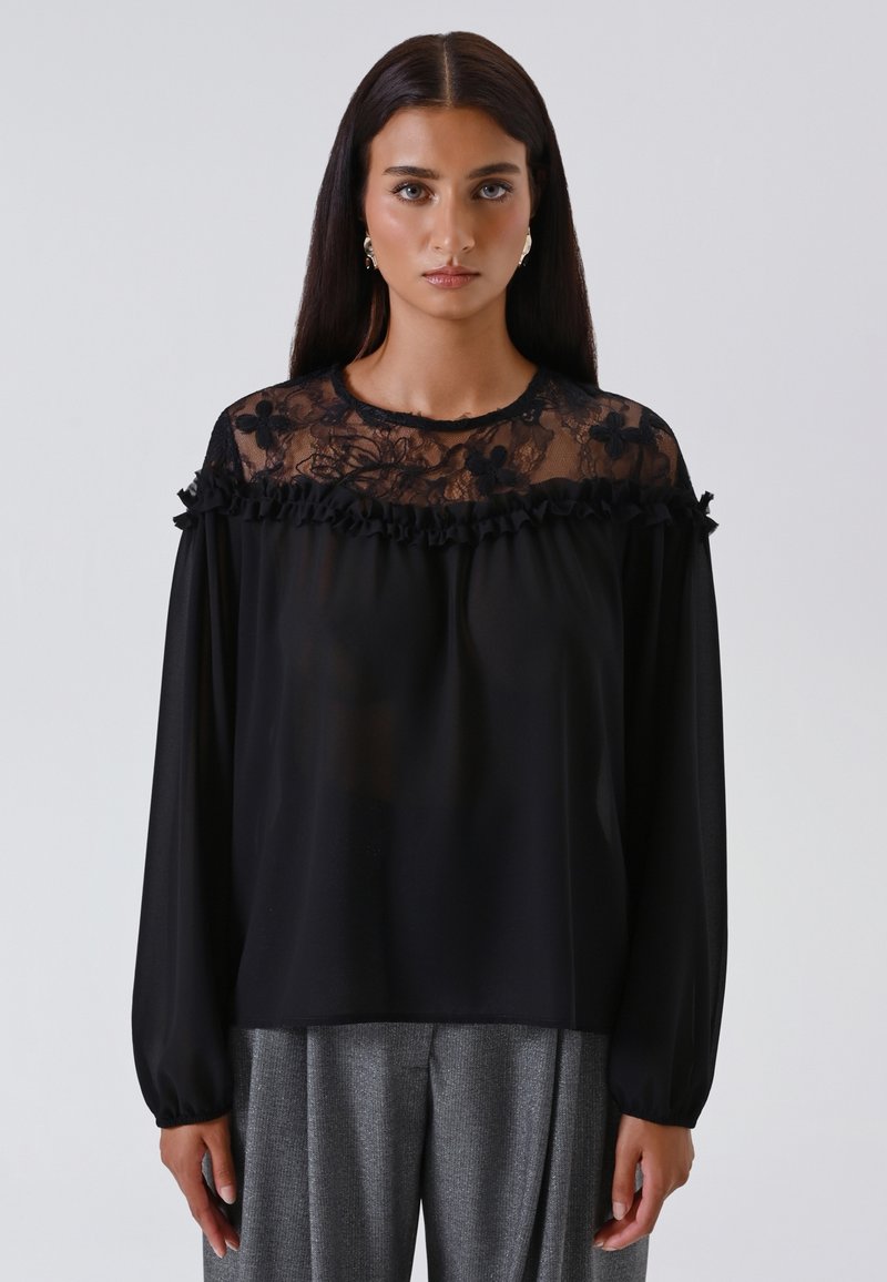 Donna con lunghi capelli scuri indossa una blusa nera trasparente con yoke in pizzo floreale e volant, abbinata a pantaloni grigi plissettati su uno sfondo semplice.