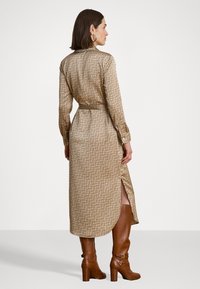 Lange, beige patroon jurk met een ceintuur in de taille, lange mouwen en zijsplitten. Gecombineerd met bruine laarzen tot de knie en hoopsieraden.