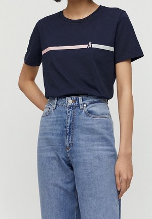 Kobieta w granatowym t-shircie z różowo-białymi poziomymi paskami oraz jasnobłękitnych, wysokich dżinsach na jednolitym tle.