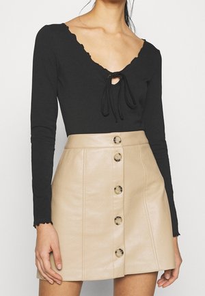 Vrouw die een zwart shirt met lange mouwen draagt met een gegolfde halslijn en een beige leren rok met knopen aan de voorkant tegen een witte achtergrond.