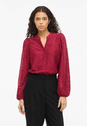 Rød teksturert bluse med bølgemønster, knapper og mandarin-krage; lange pufmermer kombinert med svarte bukser.
