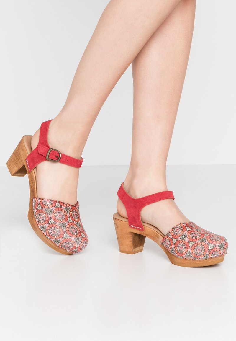 Sabots à talons en daim rouge avec un empiècement supérieur en tissu à motif floral. Comprend une bride de cheville ajustable et un talon bloc en bois. Motifs texturés sur la surface.