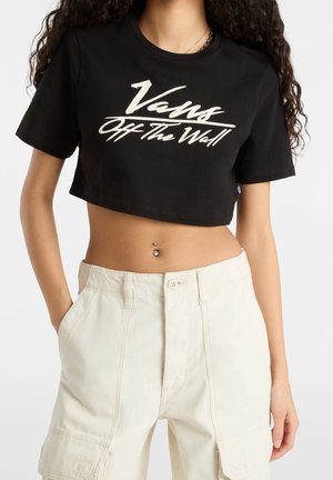 Femme portant un crop top Vans noir avec un logo blanc et un pantalon taille haute écru, main dans la poche, montrant un piercing au nombril.