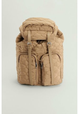 Sac à dos matelassé beige avec deux poches zippées à l'avant, des bretelles à boucle et une fermeture par cordon en haut.