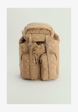 Sac à dos matelassé beige avec deux poches zippées à l'avant, des bretelles à boucle et une fermeture par cordon en haut.
