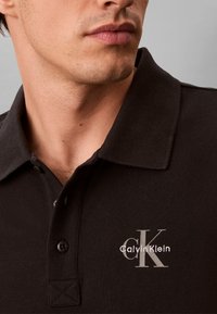 Άνδρας που φοράει μαύρο πόλο μπλουζάκι Calvin Klein με κεντημένο λογότυπο CK κοντά στον γιακά, ορατό από τον λαιμό έως το πάνω μέρος του στήθους.