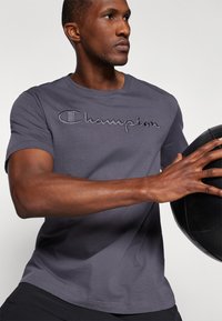 Graues Baumwoll-T-Shirt mit kurzen Ärmeln, versehen mit einem gestickten "Champion"-Logo in leicht dunklerem Grau über der Brust.