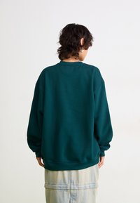 Personne avec des cheveux courts et bouclés portant des boucles d'oreilles en argent, un sweat-shirt surdimensionné de couleur vert foncé, et une jupe en denim claire, de dos.