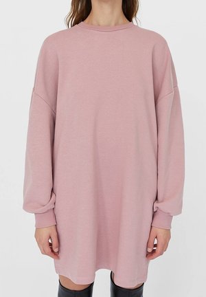 Vrouw draagt een oversized stoffige roze trui-jurk met lange mouwen, gecombineerd met zwarte kniehoge laarzen, staand tegen een effen achtergrond.