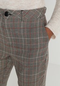 Pantalons à carreaux gris présentant des motifs pied-de-poule et à damier, avec des accents rouges. Fabriqués en tissu texturé, ils sont dotés d'une fermeture à bouton et de poches.