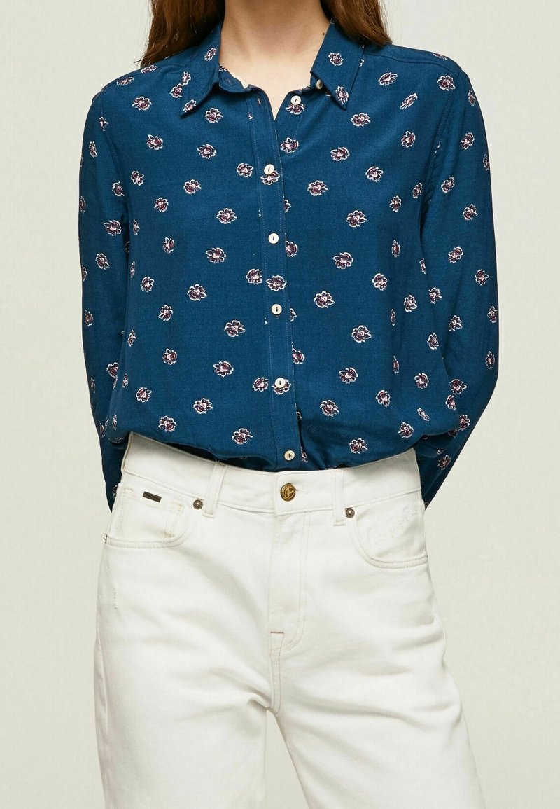 Femme portant une chemise bleue boutonnée avec un petit motif floral rentrée dans un pantalon blanc taille haute avec un bouton marron.