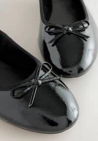 Schwarze Lackballerinas mit einer runden Spitze, die an der Vorderseite eine kleine Schleifenverzierung haben und innen mit einem weichen Stoff gefüttert sind.
