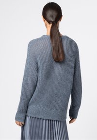 Femme aux longs cheveux noirs attachés en une queue de cheval basse, portant un pull en maille bleu ample et une jupe plissée bleue, debout devant un fond clair uni.