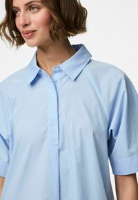 Chemise à manches courtes bleu clair avec un col classique, un plastron à boutons et des manches raglan, dotée d'une texture de tissu lisse.