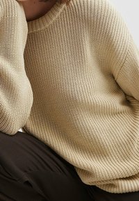 Personne portant un pull en tricot beige et un pantalon marron foncé, avec le bras reposant sur un genou levé, sur un fond uni.