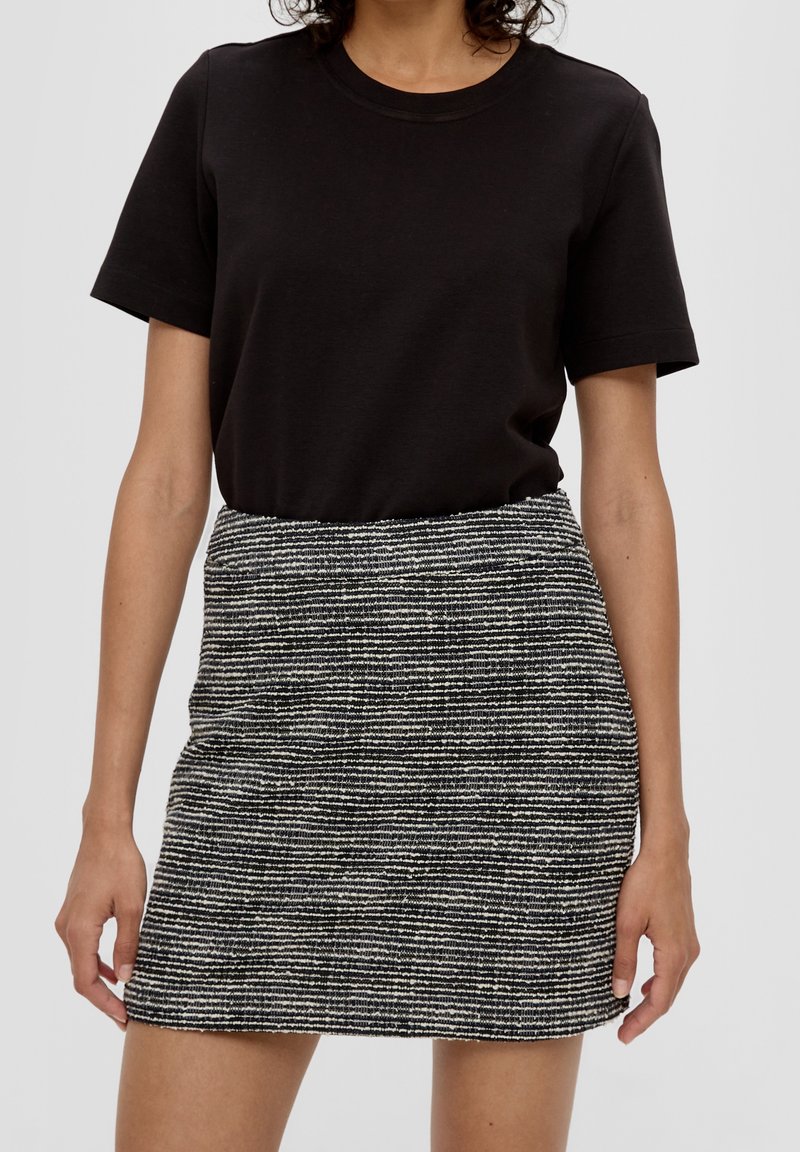 Žena v černém tričku s krátkým rukávem, zastrčeném v sukni s vysokým pasem, která je texturovaná a má černo-bílé pruhy.