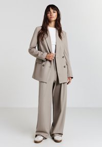 Donna con lunghi capelli castani che indossa un blazer beige doppiopetto e pantaloni larghi sopra a una maglietta bianca, con sneakers bianche, in piedi di fronte a uno sfondo neutro.