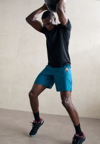 Black athletic top and teal shorts with side slits. Black sneakers with red accents. The individual is performing a weightlifting exercise.