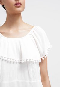 Blusa blanca con un diseño plisado, escote ancho y una capa con volantes y detalles de pompones a lo largo del dobladillo. Textura suave.