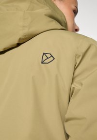 Olivgrön jacka med en tygtextur och matt finish, som visar en svart geometrisk logga på den övre ryggen.