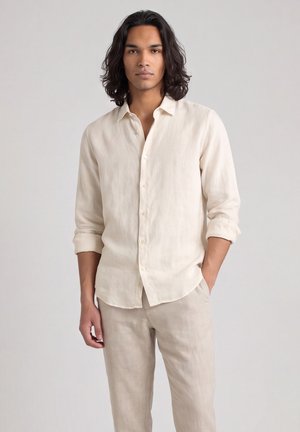 Giovane uomo con capelli scuri fino alle spalle, indossa una camicia beige con bottoni e pantaloni abbinati, in piedi con una mano in tasca.