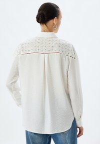 Camicia bianca a maniche lunghe con tessuto testurizzato e inserti in pizzo. Presenta polsini con bottoni e una piega posteriore per maggiore libertà di movimento.