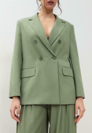 Frau trägt übergroßen, salbeigrünen, doppelt geknöpften Blazer mit passenden, weit geschnittenen Hosen vor einfarbigem Hintergrund.