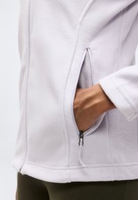 Χέρι μέσα στην τσέπη με φερμουάρ ενός ελαφρύ μπουφάν από φλις σε ανοιχτό λεβαντί χρώμα, φορεμένο πάνω από ελιάς πράσινο παντελόνι.