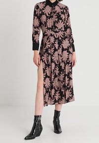 Lång svart klänning med ett rosa blommönster, knappförslutning fram och långa ärmar. Hög sprund på sidan, kombinerad med svarta kilklackboots.