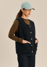 Marine fleece vest met een ronde halslijn, houten knopen en twee voorzakken, gecombineerd met een bruine lange mouwen shirt en een lichtblauwe pet.