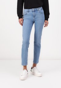 Vaqueros de mezclilla azul claro con un corte ajustado, cinco bolsillos y un ligero desgaste. Combinados con zapatillas blancas con detalles en negro.