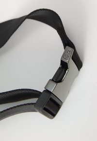 Sangle en tissu noir avec une texture tissée, présentée avec une fermeture en métal argenté brillant portant un logo gravé et une boucle en plastique réglable.