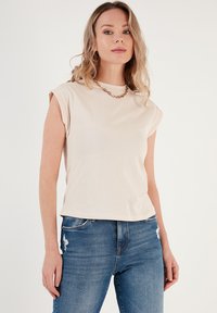 T-shirt beige a maniche corte con scollatura rotonda e risvolti, abbinata a jeans blu strappati, caratterizzata da un design minimal.