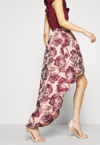 Asymetryczna sukienka z bordowym topem i różową spódnicą zdobioną dużymi bordowymi kwiatowymi wzorami; w połączeniu z cielistymi sandałami na wysokim obcasie.