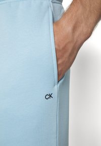Calças de algodão azul claro com um bolso lateral e um logótipo preto "CK" na bainha. Textura suave e design de ajuste relaxado.
