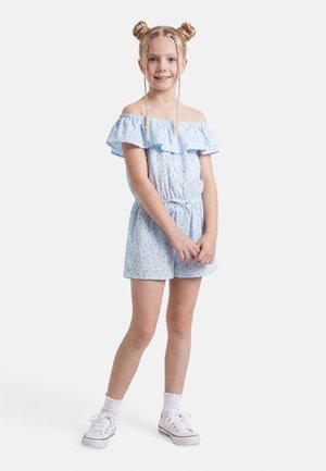 Młoda dziewczynka z blond podwójnymi kokami i warkoczami, ubrana w jasnoniebieski kwiecisty kombinezon z odkrytymi ramionami i białe trampki, stojąca i uśmiechająca się.