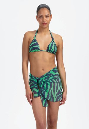 Kobieta w zielono-czarnym bikini w paski zebry z dopasowanym prześwitującym sarongiem zawiązanym w talii, stojąca na jednolitym białym tle.