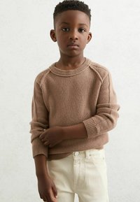 Pojke med kort krulligt hår som bär en brun stickad tröja och off-white byxor står mot en enkel ljus bakgrund.