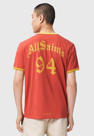 Mann trägt ein rotes und gelbes T-Shirt mit der Aufschrift „AllSaints 94“ und „MCMXCIV“ auf dem Rücken, steht vor einem schlichten hellen Hintergrund.