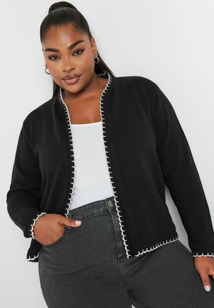 Black cardigan with white trim, textured fabric, open front design, long sleeves, paired with a white top and dark jeans.