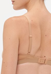 De rug van een vrouw die een verstelbare beige bh-band en haak-en-oogsluiting toont, op lichte huid met sproeten en moedervlekken.