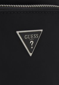 Črna najlon torba s triglavo kovinsko oznako v srebrni in črni barvi ter obliko "GUESS USA" in vprašaj. Vidne podrobnosti z zadrgo.