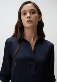 Navyblauwe satijnen blouse met een knoopsluiting aan de voorkant, lange mouwen en een scherpe kraag. Gladde textuur en glanzende afwerking.